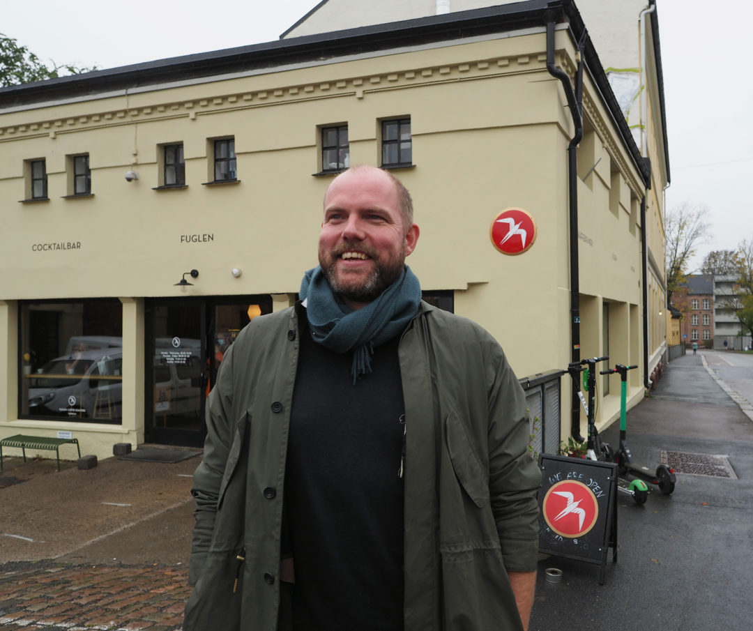 En mann (Einar) står utenfor kafeen Fuglen i Gamlebyen og ser til siden. Han smiler og har på seg jakke og skjerf.